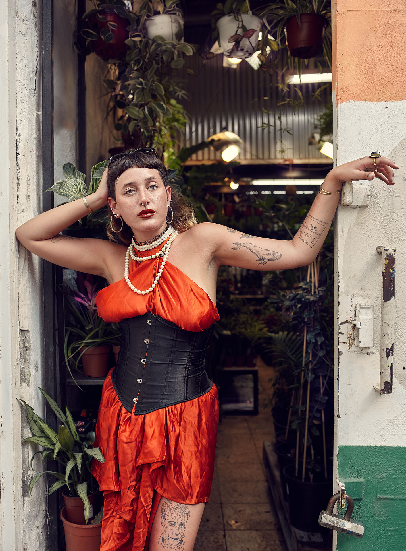 Person mit dunklen langen Haaren und kurzen Pony schaut direkt in die Kamera. Die Person trägt ein orangenes Kleid welches mit einem Korsett zusammen gehalten wird. Die Person lehnt an einer Tür oder Durchgang zu einem Geschäft. Im Hintergrund befinden sich Pflanzen.